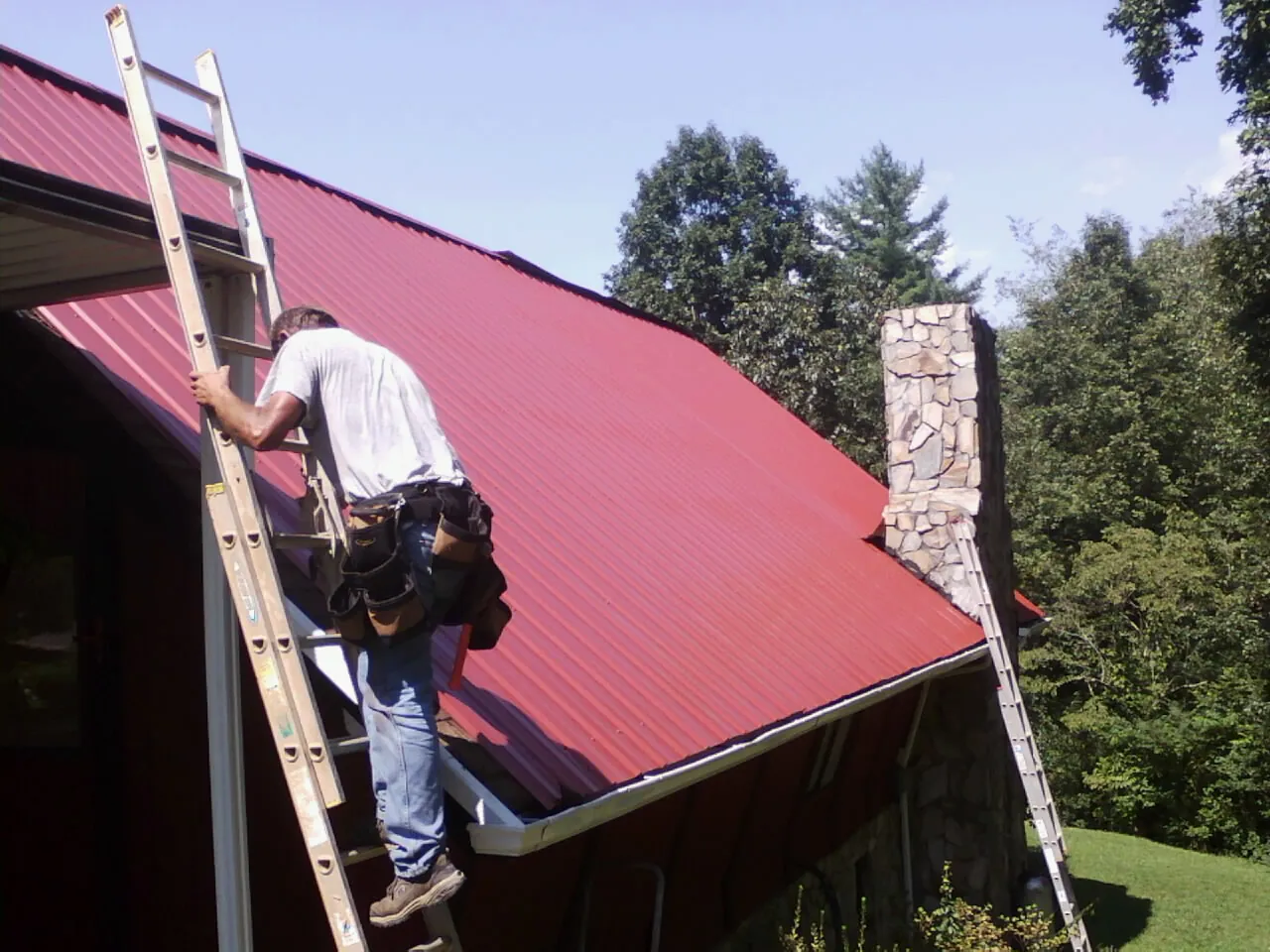 Roofer on a ladder beside a red metal roof and stone chimney — Soffit & Fascia Repair by Roofing Brothers in New Paltz